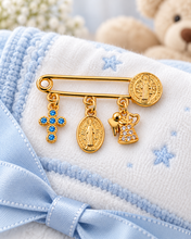 Blue cross and Saint Benedict baby blessing pin with Virgin Mary medal and angel charm on a blue and white blanket